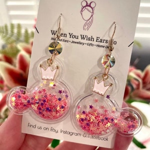 May include: Pink and gold glitter filled Mickey Mouse shaped earrings with a crown on top. The earrings are on a white card with the text "When You Wish Ears" and "Find us on Etsy, Instagram & Facebook".