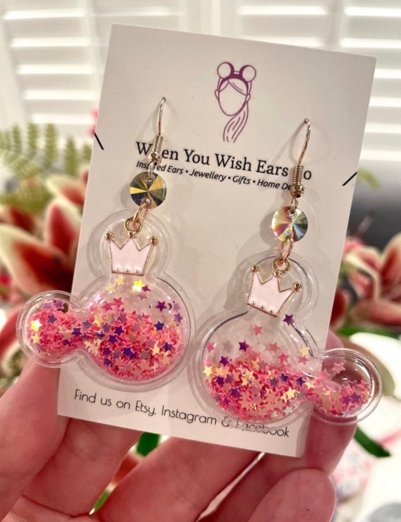 May include: Pink and gold glitter filled Mickey Mouse shaped earrings with a crown on top. The earrings are on a white card with the text "When You Wish Ears" and "Find us on Etsy, Instagram & Facebook".
