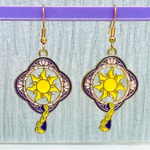 May include: A pair of gold-coloured dangle earrings. Each earring features a sun design with yellow, pink, and purple enamel. The sun is surrounded by a decorative frame and a dangling braid. The earrings are suspended from gold-coloured ear wires.