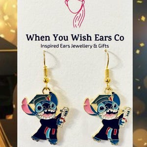 May include: A pair of gold dangle earrings featuring a blue Stitch character wearing a graduation cap and gown, holding a diploma. The earrings are from When You Wish Ears Co, Inspired Ears Jewellery & Gifts. The text "On Etsy, Instagram, Facebook" is also visible.
