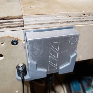 May include: A gray, rectangular device with a geometric design is mounted on a wooden surface. The device is held in place by a gray bracket. A black cable and a small black cylindrical object are connected to the device.