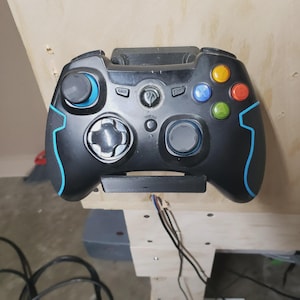 May include: Black gaming controller with blue accents and multi-colored buttons. The controller has a directional pad, two analog sticks, and several buttons labeled "Back", "Start", and "Turbo". The controller is mounted on a wooden surface.
