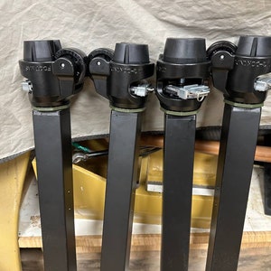 May include: Four black metal rolling bases with wheels and a locking mechanism. Each base has a black square column and a black top with a wheel on each side.