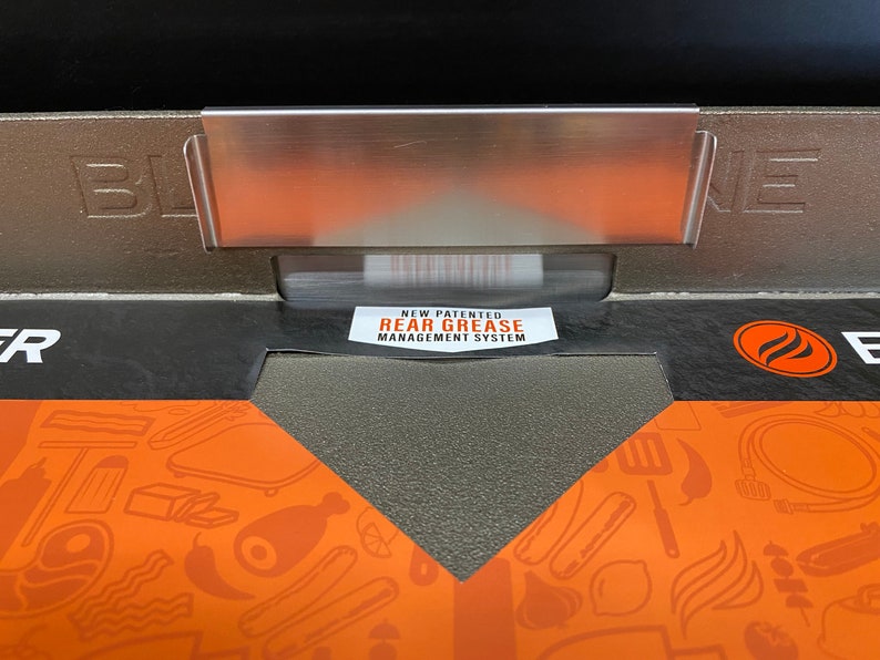 May include: Close-up of a black grill with a stainless steel grease management system. The grill has a black and orange design with the words "Blaze" and "New Patented Rear Grease Management System" printed on it.