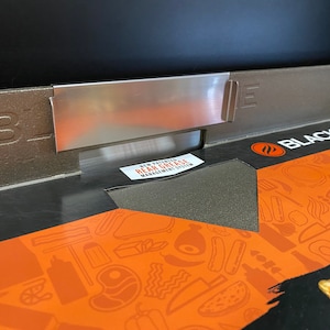 May include: Close-up of a black Blackstone griddle with a stainless steel grease management system. The griddle has a black and orange patterned surface with the text "Blackstone" printed on it.