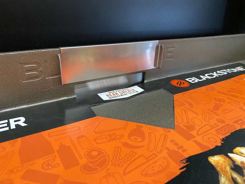 May include: Close-up of a black Blackstone griddle with a stainless steel grease management system. The griddle has a black and orange patterned surface with the text "Blackstone" printed on it.