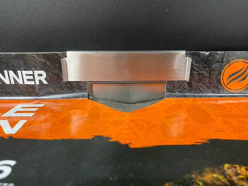 May include: A close-up of a silver metal sign holder with a black background. The sign holder is attached to a package with the word "RUNNER" printed on it. The package has an orange background with a pattern of various icons.