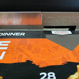 May include: Close-up of a black Blackstone grill with a stainless steel side shelf. The grill has a new patented rear grease management system. The grill is on a box with a colorful design featuring various grilling tools and food items. The box also has the text "Each Dinner" and "28" printed on it.