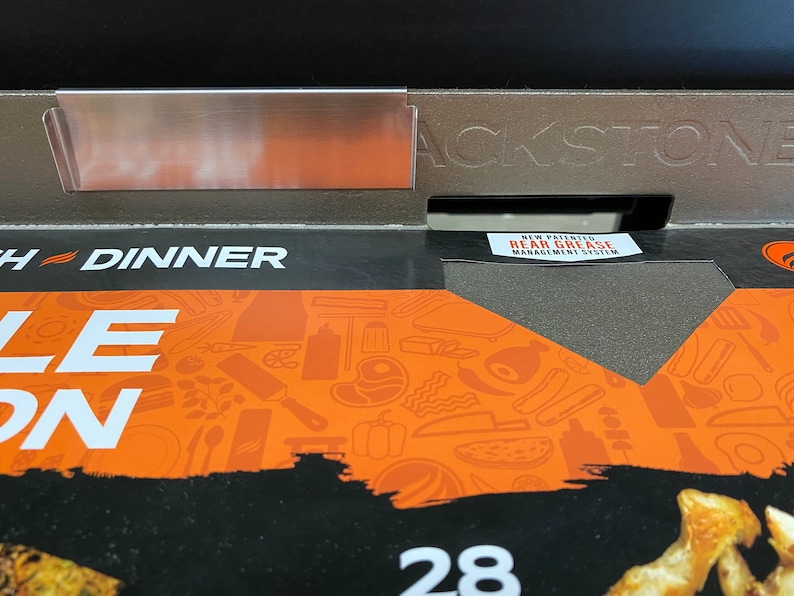 May include: Close-up of a black Blackstone grill with a stainless steel side shelf. The grill has a new patented rear grease management system. The grill is on a box with a colorful design featuring various grilling tools and food items. The box also has the text "Each Dinner" and "28" printed on it.