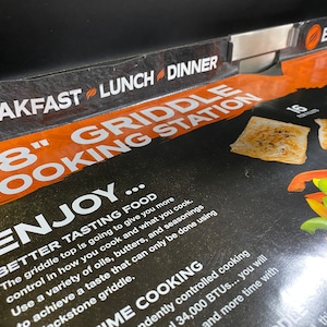 May include: A close-up of a black and orange 28-inch griddle cooking station with the text "BREAKFAST LUNCH DINNER" and "ENJOY... BETTER TASTING FOOD" on the front. The griddle is shown with cooked food, including peppers and fish.