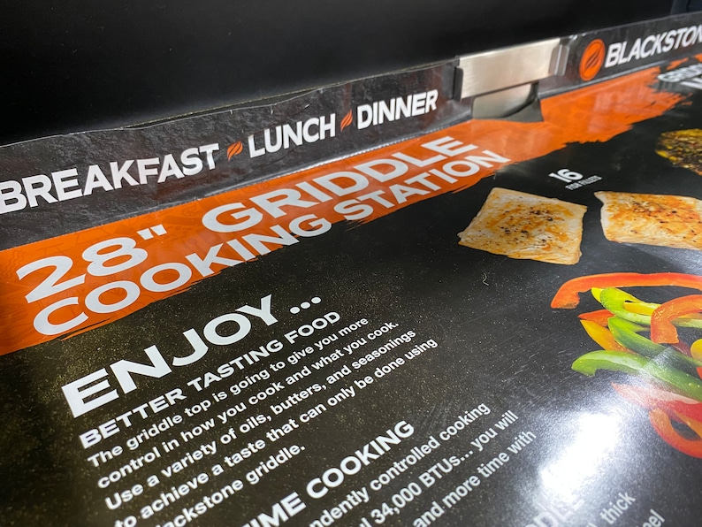 May include: A close-up of a black and orange 28-inch griddle cooking station with the text "BREAKFAST LUNCH DINNER" and "ENJOY... BETTER TASTING FOOD" on the front. The griddle is shown with cooked food, including peppers and fish.