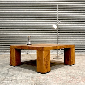 May include: A large, square wooden coffee table with a natural wood grain finish. The table has four thick, rectangular legs. A silver metal lamp and a decorative glass object sit on the table.