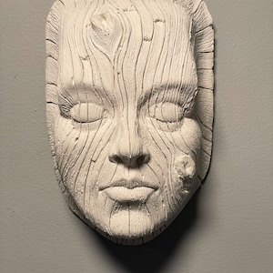 May include: A white, textured wall sculpture of a human face with closed eyes. The face is carved with a wood grain pattern, giving it a rustic appearance. The sculpture is mounted on a gray wall.