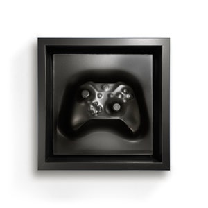 Puede incluir: Un mando de videojuegos negro en una caja negra con un contorno en relieve del mando.