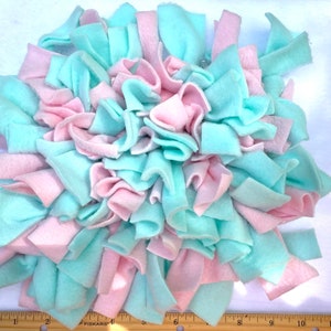 May include: A handmade, round pet bed made of soft fleece strips in alternating pastel pink and light blue colors. The strips are knotted together, creating a textured, plush surface. The bed is designed for comfort and is suitable for small pets.