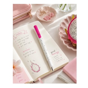 May include: An open notebook with handwritten notes, a drawing of a dragon fruit, and the text "notes", "REMINDER", and "good energy". A white fountain pen with a pink cap and black dots rests on the page. Nearby are pink stationery items.