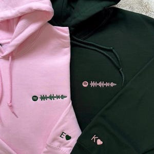May include: Two hoodies, one pink and one dark green, are displayed. Each hoodie features a Spotify code and a small embroidered initial with a heart on the sleeve. The pink hoodie has a matching pink drawstring, while the green hoodie has a green drawstring.