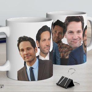 May include: Two white ceramic mugs with a group of men's faces printed on them. The mugs are on a wooden table with a laptop, a pencil, and a plant in the background.