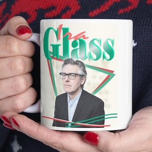 May include: White ceramic mug with a green and red graphic design featuring a man wearing glasses and the word "Glass".