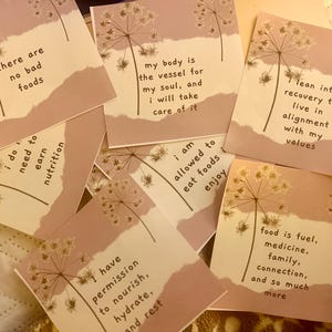May include: A set of ten pink and white affirmation cards with floral designs and inspirational messages. The cards include phrases like "There are no bad foods", "I am allowed to eat foods and enjoy", and "Food is fuel, medicine, family, connection, and so much more".