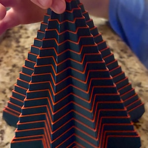 May include: A dark blue and orange geometric pyramid-shaped toy. The toy has a stepped design with orange lines outlining each step. A small red piece sits on top. The toy is held by a person.