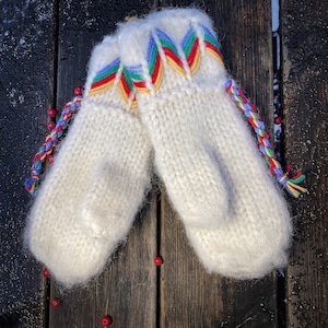 Puede incluir: Un par de guantes de punto blancos con ribetes trenzados de colores. Los guantes están sobre una superficie de madera.