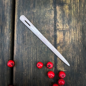 May include: A white wooden needle with a small hole in the centre, lying on a dark brown wooden surface with red berries scattered around it.