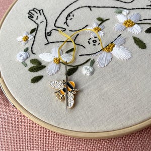 May include: Embroidery hoop with a floral design and a bee needle minder. The embroidery features a line drawing of a figure surrounded by white flowers with yellow centers and green leaves. A yellow thread and needle are visible.