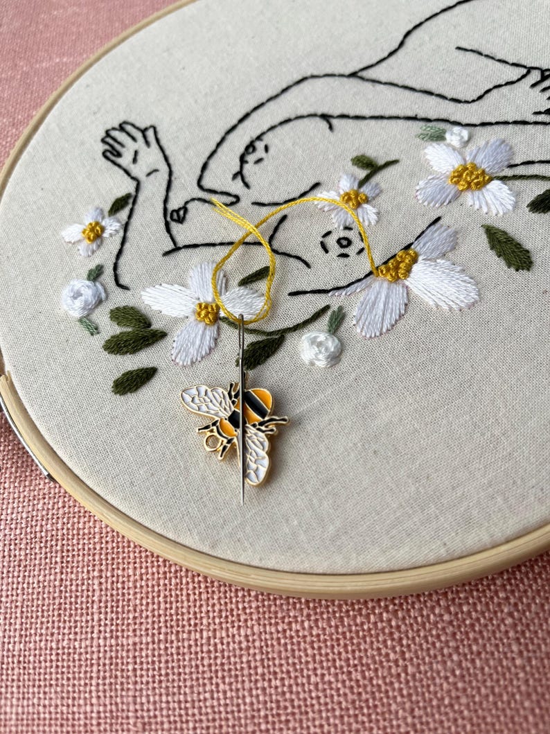 May include: Embroidery hoop with a floral design and a bee needle minder. The embroidery features a line drawing of a figure surrounded by white flowers with yellow centers and green leaves. A yellow thread and needle are visible.