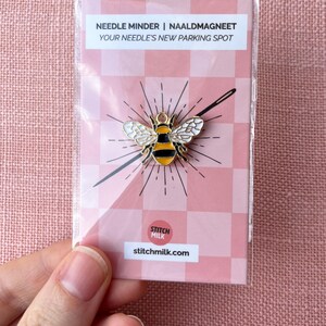 May include: A bee-shaped needle minder with black and yellow stripes and white wings, packaged on a pink and white checkered card. The packaging reads "Needle Minder | Naaldmagneet" and "Your Needle's New Parking Spot."