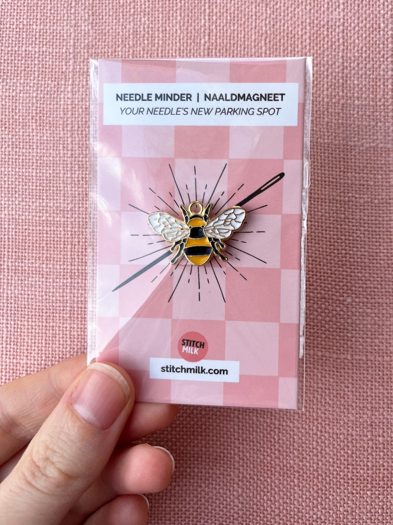 May include: A bee-shaped needle minder with black and yellow stripes and white wings, packaged on a pink and white checkered card. The packaging reads "Needle Minder | Naaldmagneet" and "Your Needle's New Parking Spot."