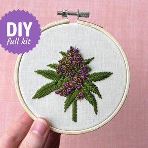 Op de afbeelding: Een doe-het-zelf borduurpakket met een cannabis knop ontwerp. De borduursel zit in een houten ring, met groene bladeren en paarse knoppen. De tekst "DIY full kit" staat in een paarse ster.