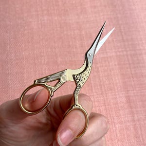 Può includere: Forbici da ricamo in metallo dorato a forma di gru con un design ad ala testurizzata e lame color argento. I manici formano il corpo e le zampe dell'uccello, mentre le lame appuntite si estendono dalla testa con una piccola vite di fissaggio.