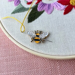 May include: A close-up of an embroidery hoop with a floral design. The embroidery features purple, red, and yellow flowers with green leaves. A bee-shaped needle minder is attached to the fabric with a needle and yellow thread.