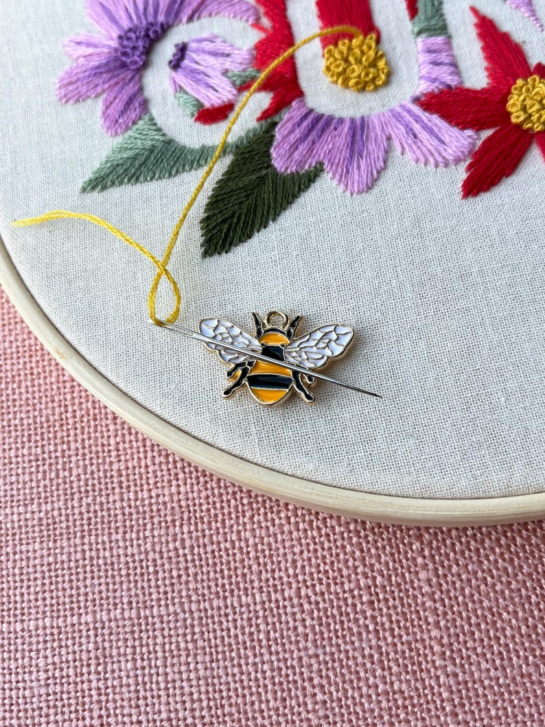 May include: A close-up of an embroidery hoop with a floral design. The embroidery features purple, red, and yellow flowers with green leaves. A bee-shaped needle minder is attached to the fabric with a needle and yellow thread.