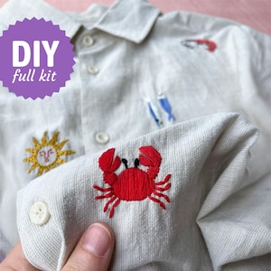 May include: Cream-coloured button-down shirt with embroidered details. A red crab is embroidered on the cuff, with a yellow sun and small blue and red sea creatures. The text "DIY full kit" is in a purple starburst.