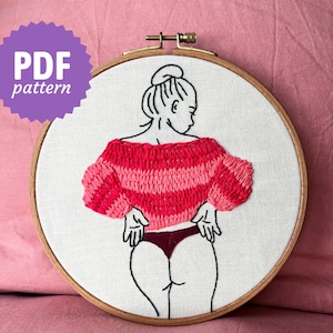 Può includere: Arte ricamato su un cerchio con un disegno a linea di una persona che indossa un maglione a righe rosa e rosso e un fondo bikini rosso scuro. L'opera d'arte è su uno sfondo bianco ed è incorniciata in un cerchio di legno.