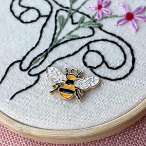 May include: A gold-colored bee charm with black and yellow stripes and white wings rests on a canvas embroidery hoop. The embroidery features black swirls, green stems, and pink flowers with red centers. The hoop is made of light-colored wood.
