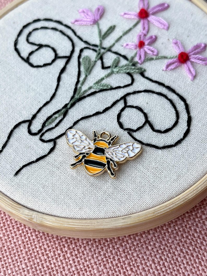 May include: A gold-colored bee charm with black and yellow stripes and white wings rests on a canvas embroidery hoop. The embroidery features black swirls, green stems, and pink flowers with red centers. The hoop is made of light-colored wood.