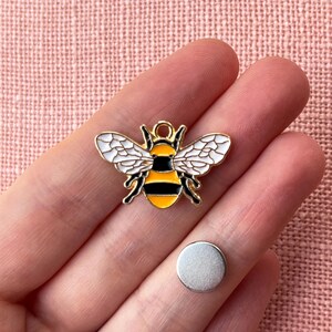 May include: A gold-colored bee charm with black and yellow stripes, white wings, and a small silver disc. The bee charm is a decorative item, possibly for jewelry or crafts.