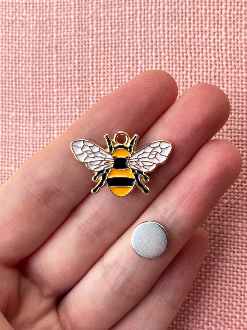 May include: A gold-colored bee charm with black and yellow stripes, white wings, and a small silver disc. The bee charm is a decorative item, possibly for jewelry or crafts.