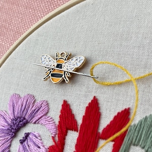 May include: Close-up of embroidery hoop with a bee-shaped pin and needle. The needle is threaded with yellow thread, and the fabric features embroidered flowers in purple, red, and green. The background is a pink fabric.