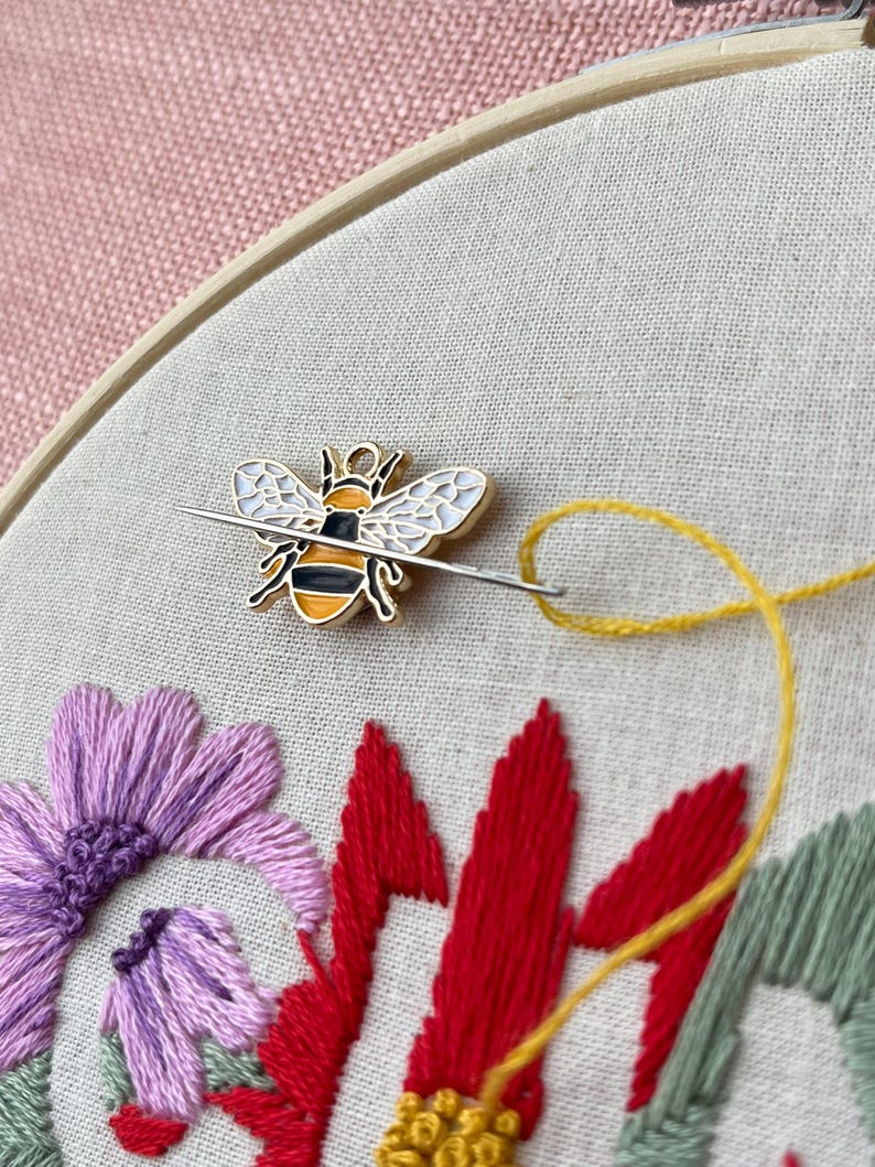 May include: Close-up of embroidery hoop with a bee-shaped pin and needle. The needle is threaded with yellow thread, and the fabric features embroidered flowers in purple, red, and green. The background is a pink fabric.