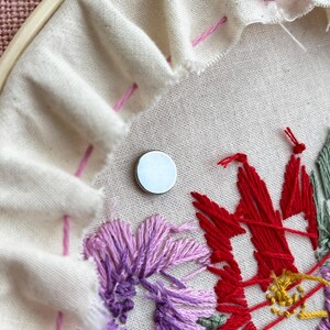May include: Close-up of an embroidery project in progress. A white fabric is stretched within a wooden hoop, featuring a partially completed floral design in shades of red, purple, and green. A small, silver magnet is placed on the fabric.