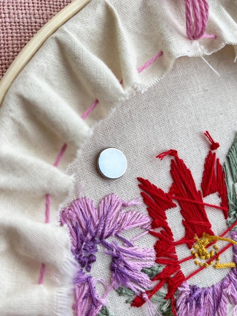 May include: Close-up of an embroidery project in progress. A white fabric is stretched within a wooden hoop, featuring a partially completed floral design in shades of red, purple, and green. A small, silver magnet is placed on the fabric.