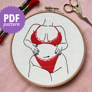Könnte beinhalten: Stickerei einer Person in einem roten Spitzen-Dessous-Set auf einem weißen Stoffhintergrund. Das Design ist in einem Stickrahmen.