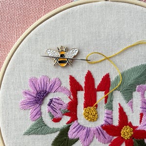 May include: An embroidery hoop with a floral design and the word "SUN" stitched in colorful thread. A bee-shaped pin with black, yellow, and white enamel is attached to the fabric with a needle and yellow thread. The background is a light pink fabric.