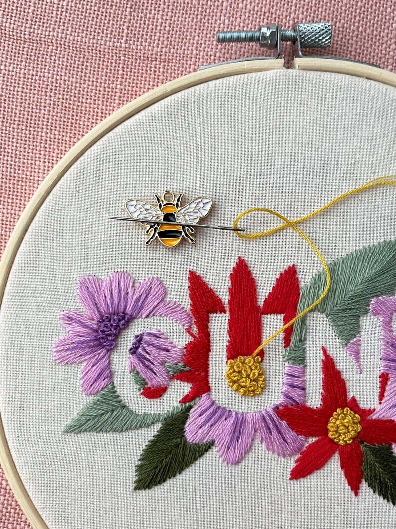 May include: An embroidery hoop with a floral design and the word "SUN" stitched in colorful thread. A bee-shaped pin with black, yellow, and white enamel is attached to the fabric with a needle and yellow thread. The background is a light pink fabric.