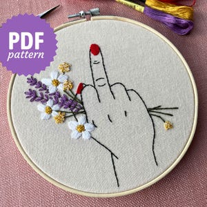May include: Embroidered hoop art featuring a hand giving the middle finger with red nail polish, surrounded by a bouquet of white daisies and purple lavender.