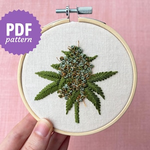 May include: A circular wooden embroidery hoop containing a piece of off-white fabric with a hand-stitched design of a green cannabis bud and leaves. A purple starburst graphic in the upper left corner contains the white text PDF pattern.